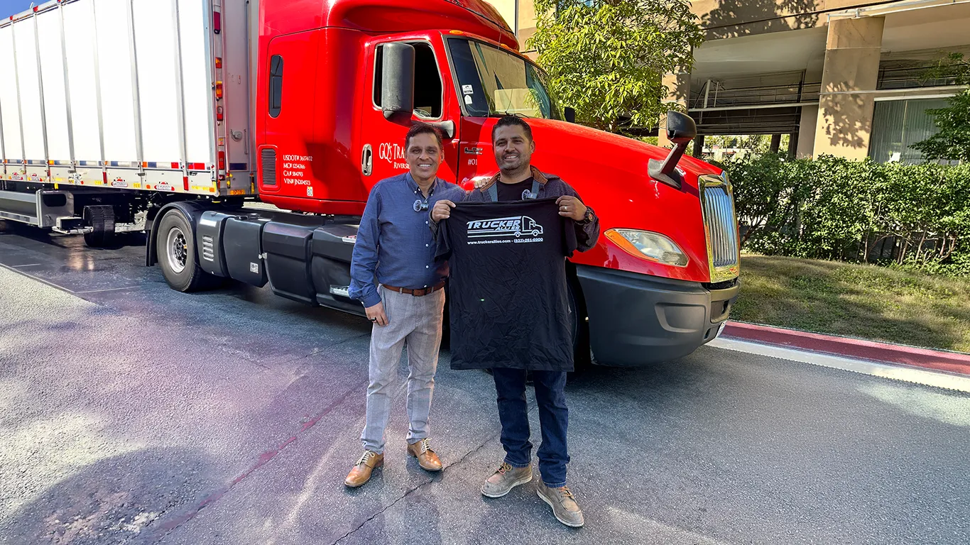 Jorge H., a Trucker Allies client, standing with a member of the Allies team in front of a red GQ’s Transport semi truck and holding up a Trucker Allies shirt.