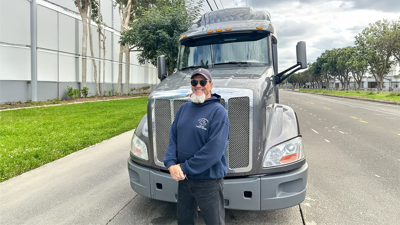 Larry H., a Trucker Allies client, in sunglasses and a navy hoodie, standing in front of the chrome grille of a grey semi truck.
